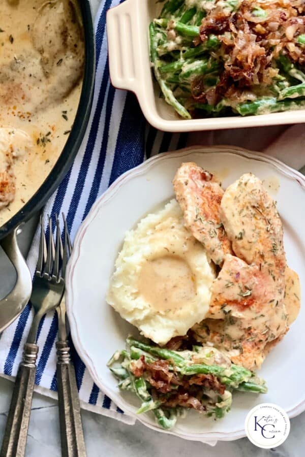 Plate with mashed potatoes, green bean casserole, and turkey breast cutlets with 2 forks, and green bean casserole in the background.