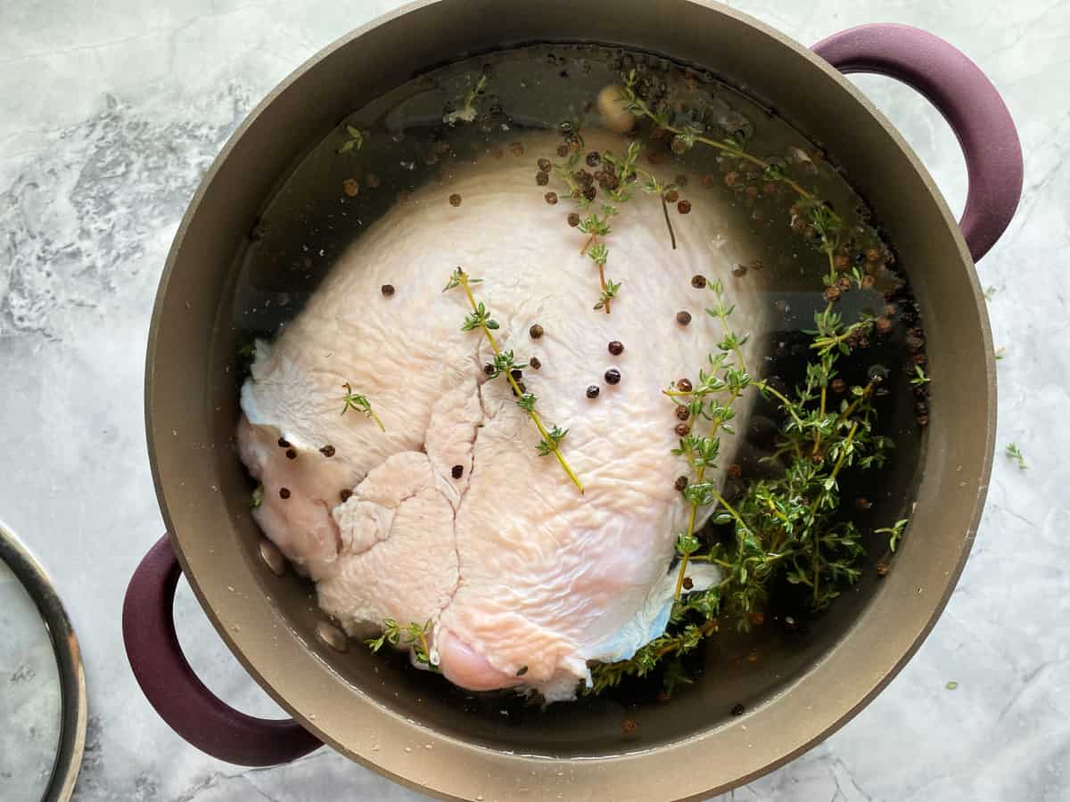 Brown pot on a marble countertop filled with liquid, fresh thyme, black peppercorns, and raw turkey breast.