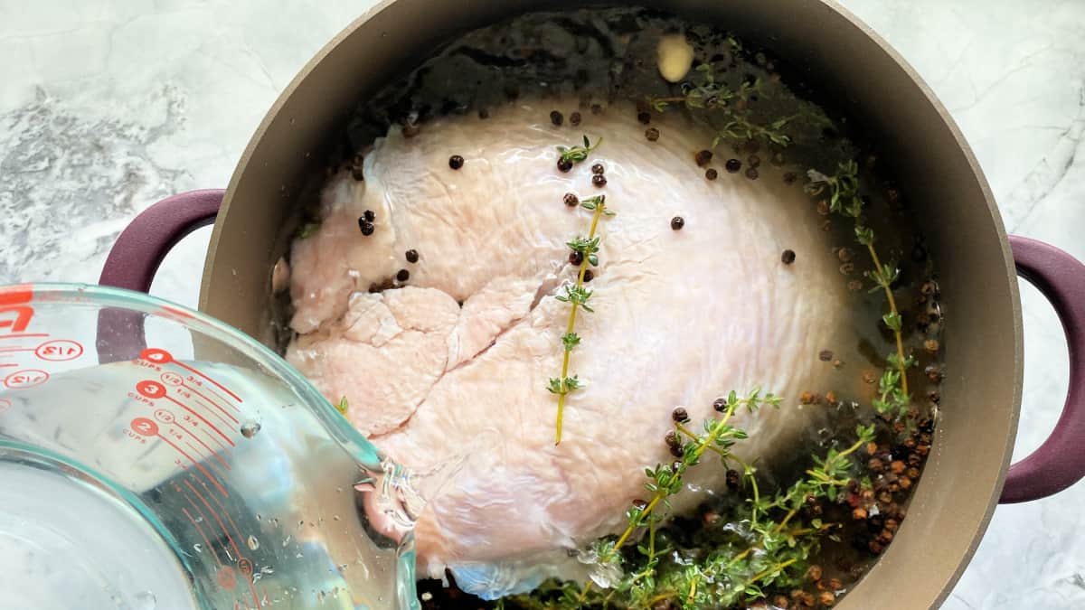 Brown pot on a marble countertop with a raw turkey, black peppercorns, and thyme in the pot with a glass measuring cup pouring in water.