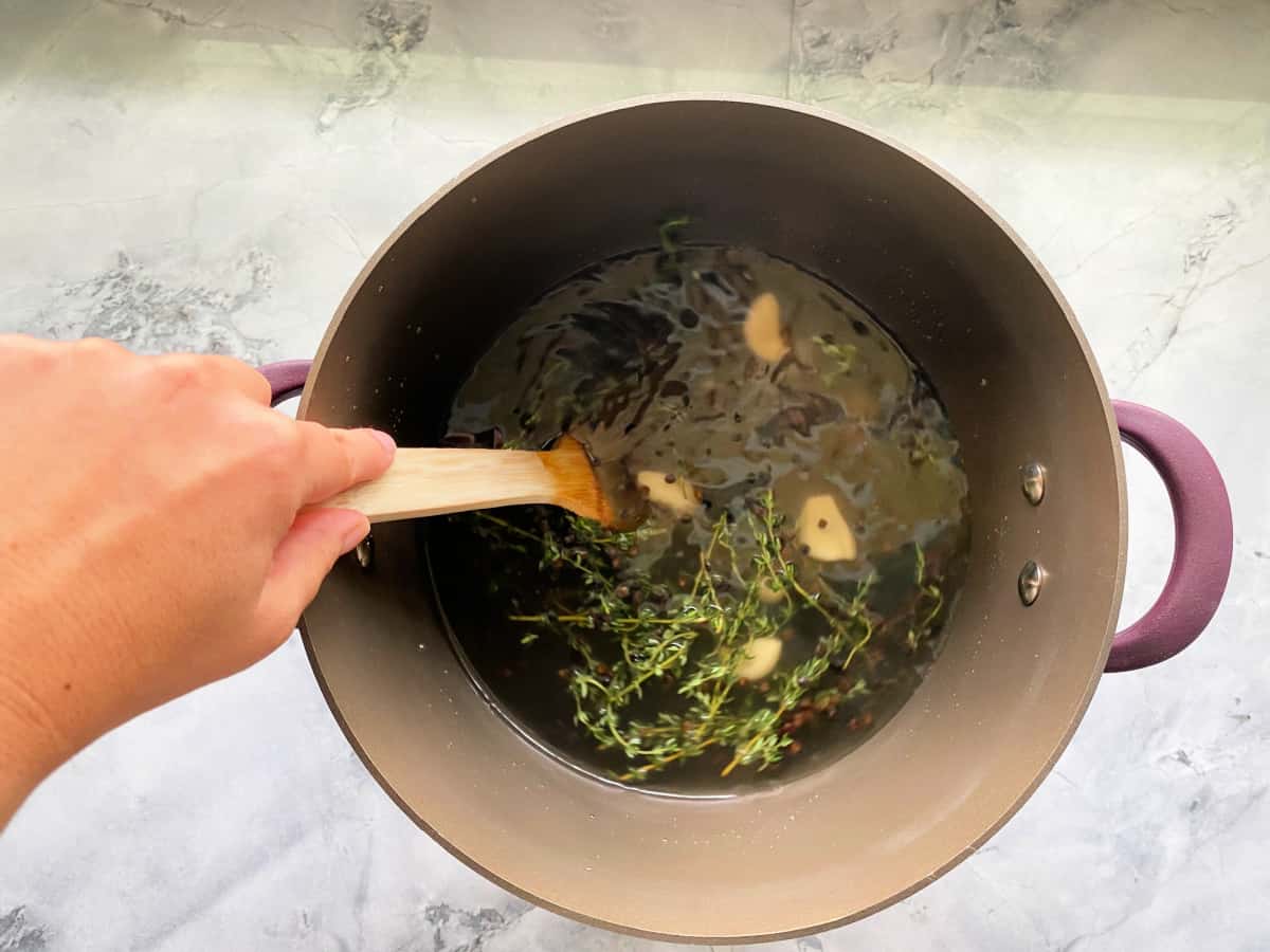 Brown pot on a marble countertop with a hand holding a wood spoon stirring liquid with fresh thyme and garlic cloves.