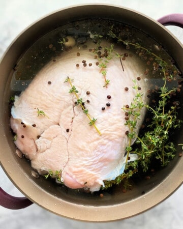 Brown pot on a marble countertop with a raw turkey breast and spices inside it.