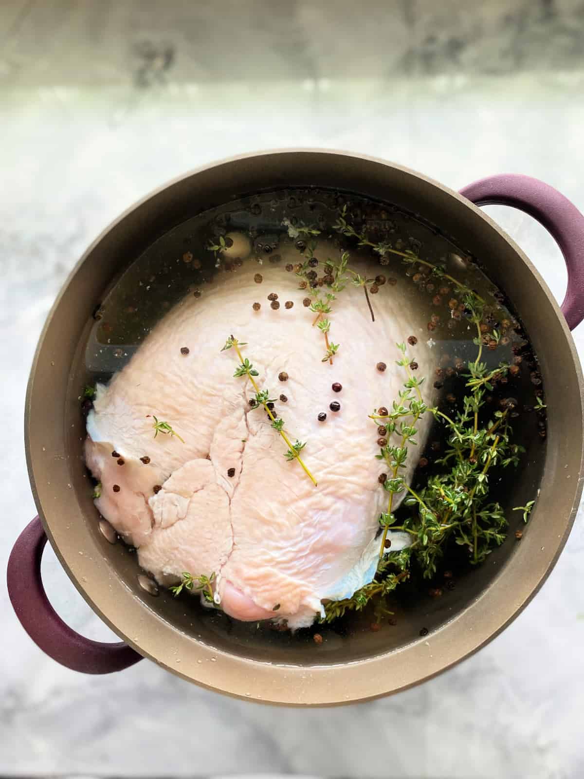 Brown pot with a turkey peppercorns, and fresh thyme in water on a marble countertop.