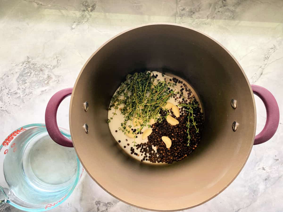 Brown pot on a marble countertop filled with salt, garlic cloves, pepercorn, and thyme with water on the side.