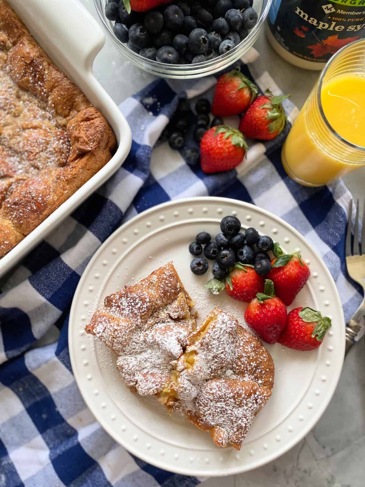 White plate filled with two slices of baked french toast with berries on a blue and white checkered napkin with orange juice next to it.