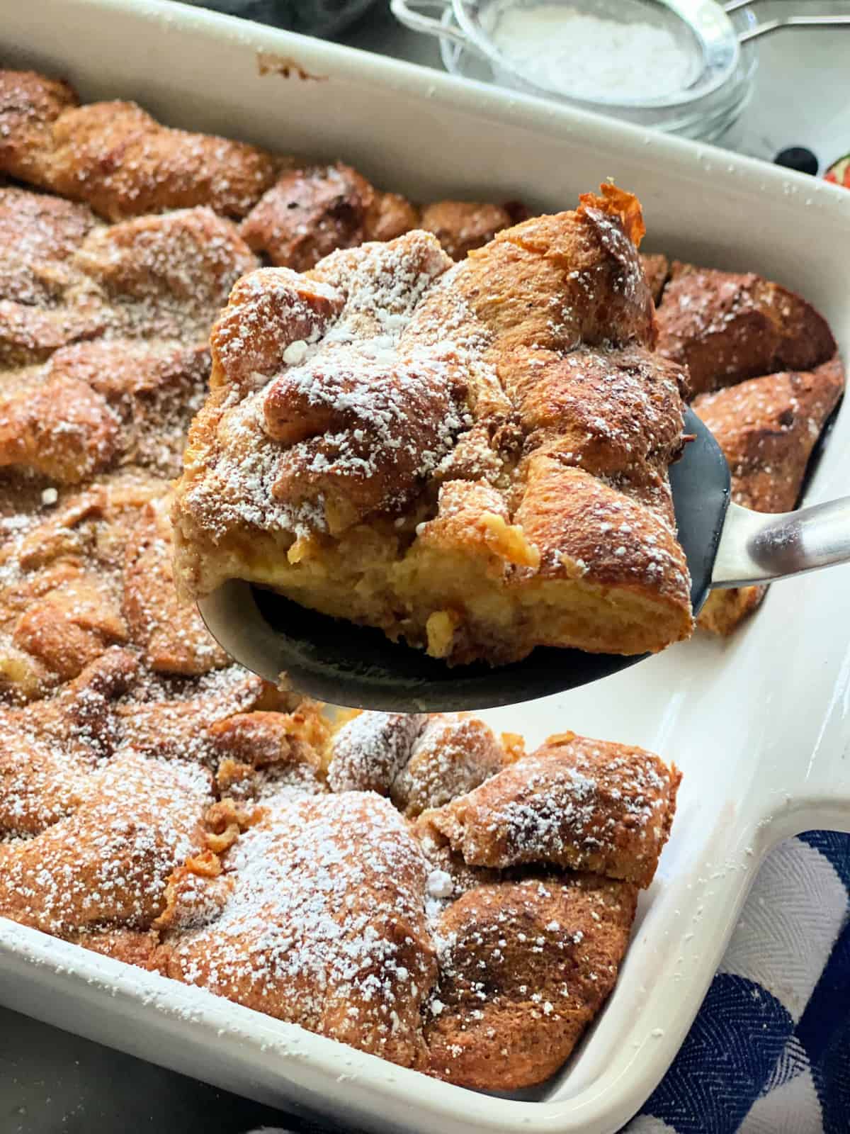 White baking dish filled with baked french toast with a black spatula holding a slice over the baking dish.