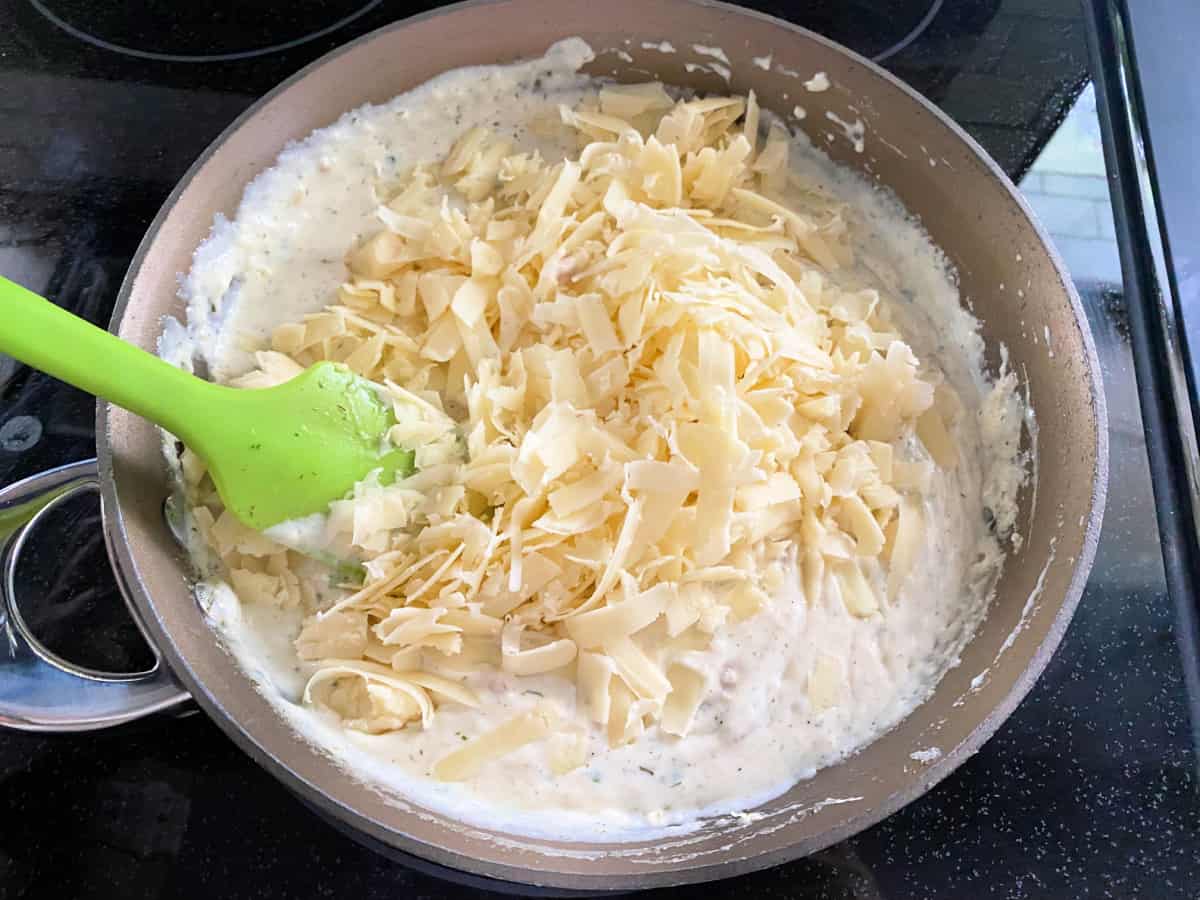 Shredded cheese with cream sauce in a saucepan on the stove top being mixed with a green spatula.