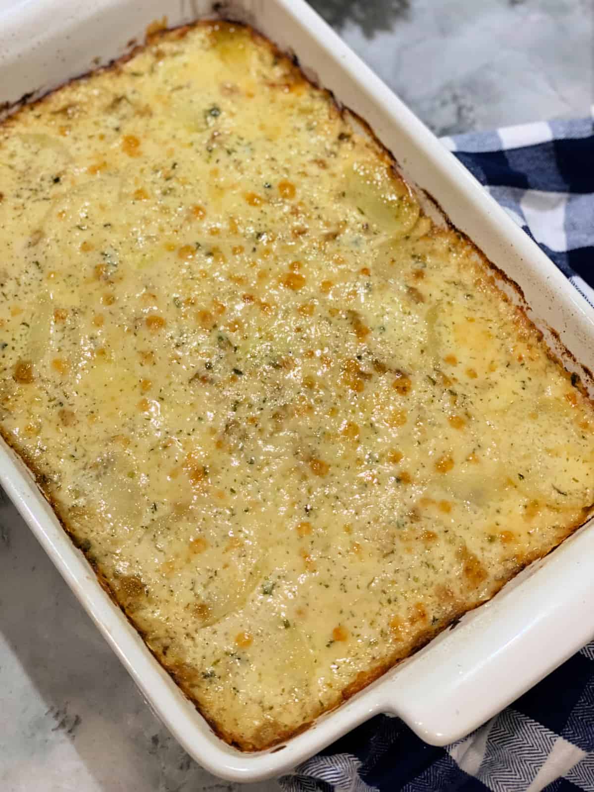 White rectangular casserole dish with a cheesy casserole baked in it sitting on a blue and white checkered cloth and marble countertop.