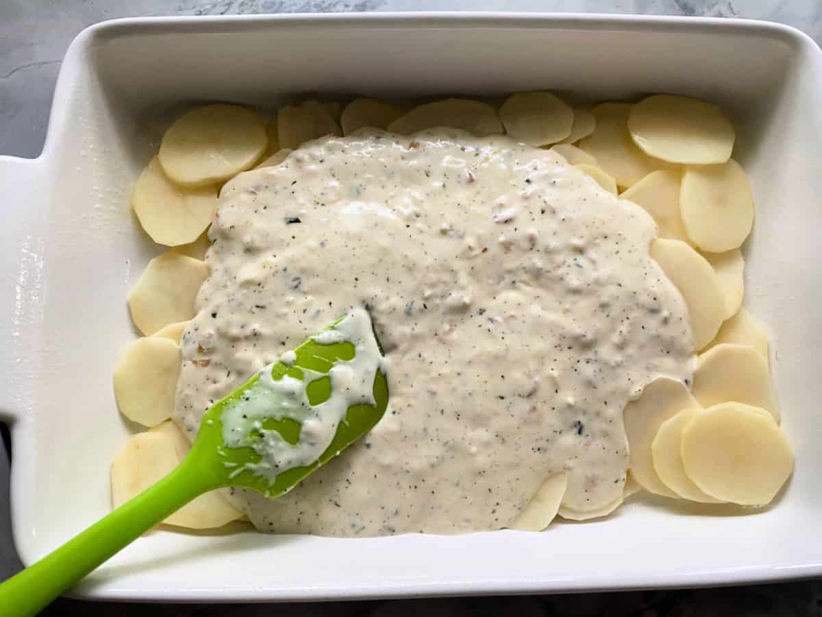 Marble countertop with a white casserole dish filled with sliced potatoes getting filled with cream sauce with green spatula.