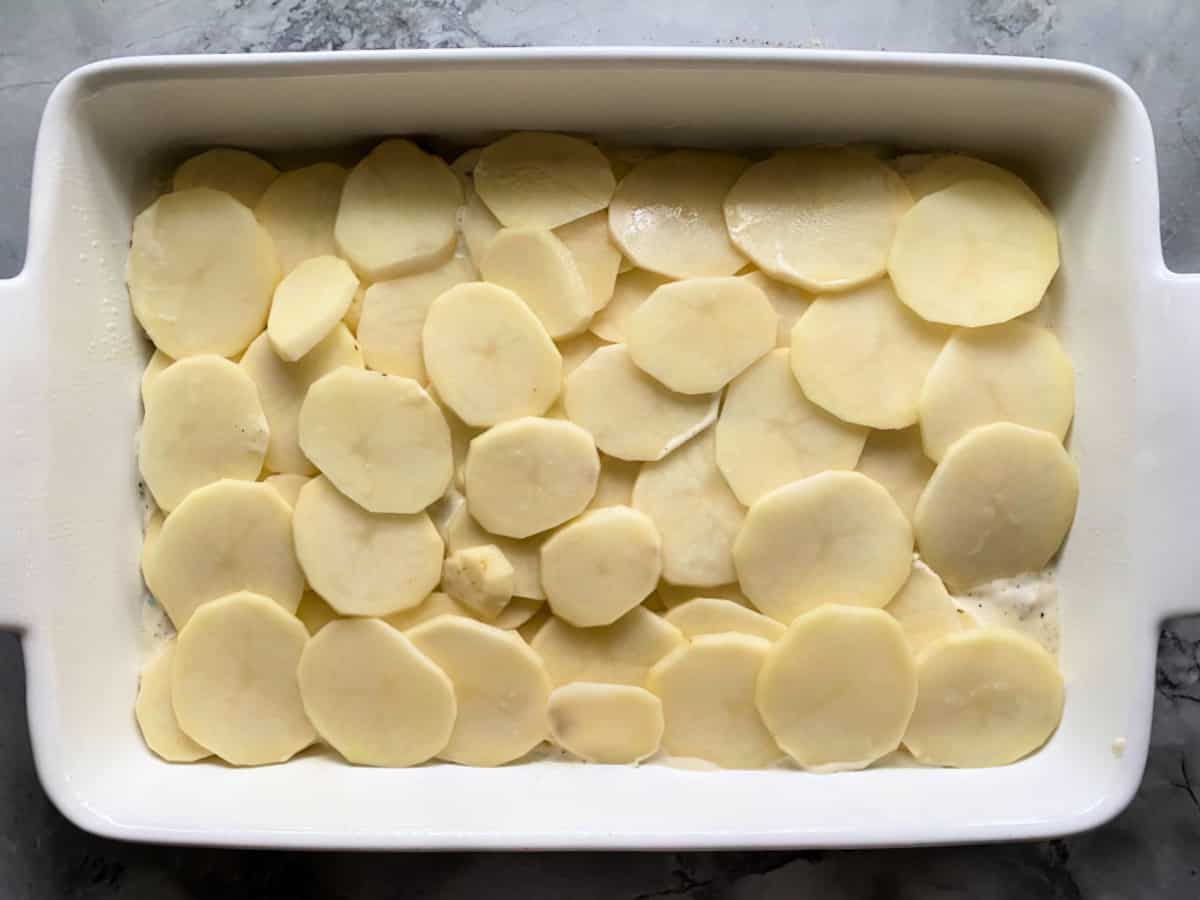 Marble countertop with a white casserole dish filled with sliced potatoes.