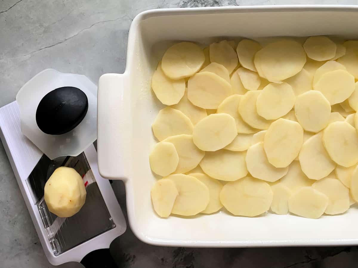 Marble countertop with a white casserole dish filled with slice potatoes with potato slicer sitting on the counter.