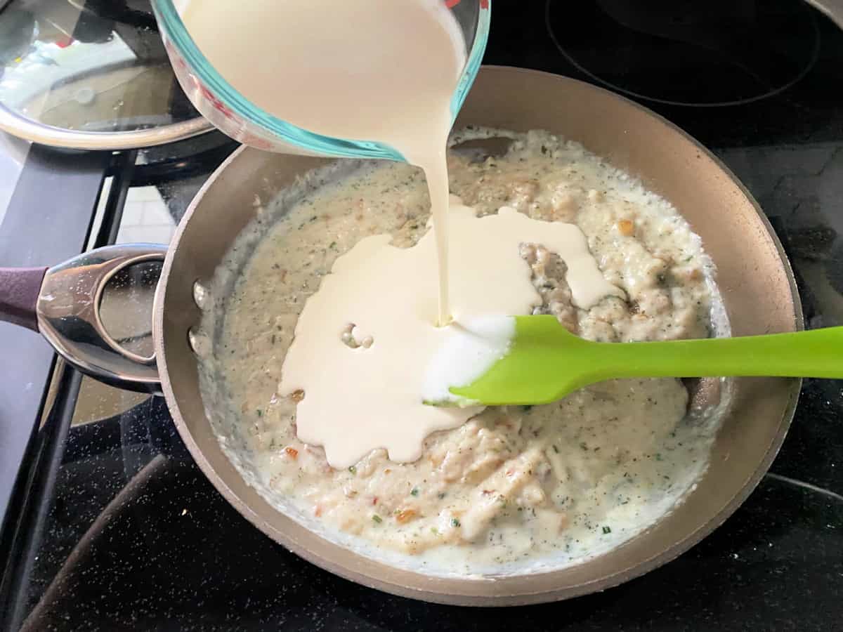 Heavy cream being added to the cream sauce in the saucepan on top of the stove top with a green spatula.