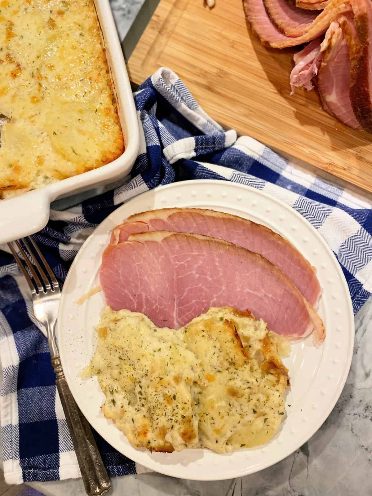 Cheesy Scalloped Potatoes with sliced ham on a white dish sitting on top of a plaid blue and white kitchen towel.