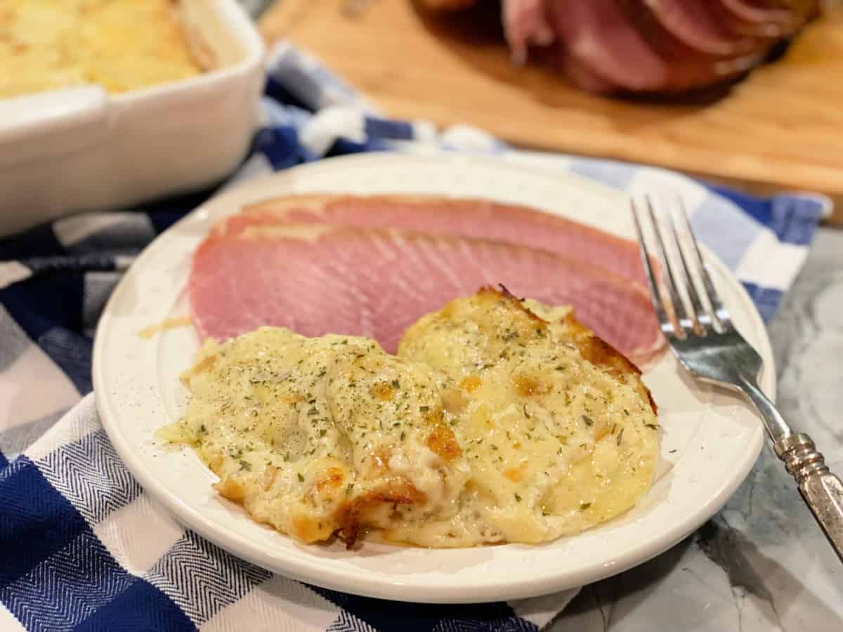 Side view of Cheesy Scalloped Potatoes with sliced ham on a white dish sitting on top of a plaid blue and white kitchen towel.