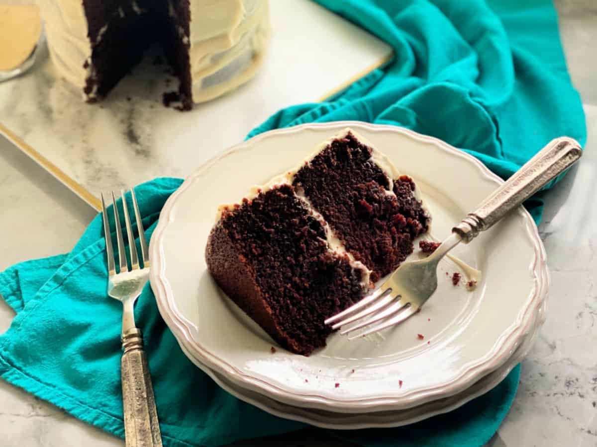 Slice of chocolate cake with cream cheese frosting on a white plate with blue table runner.
