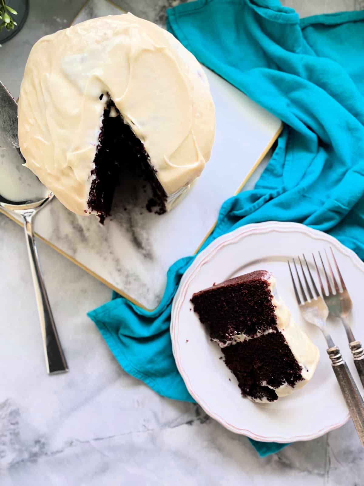 Slice of Chocolate Cake with Cream Cheese Frosting with entire cake on a marble countertop.