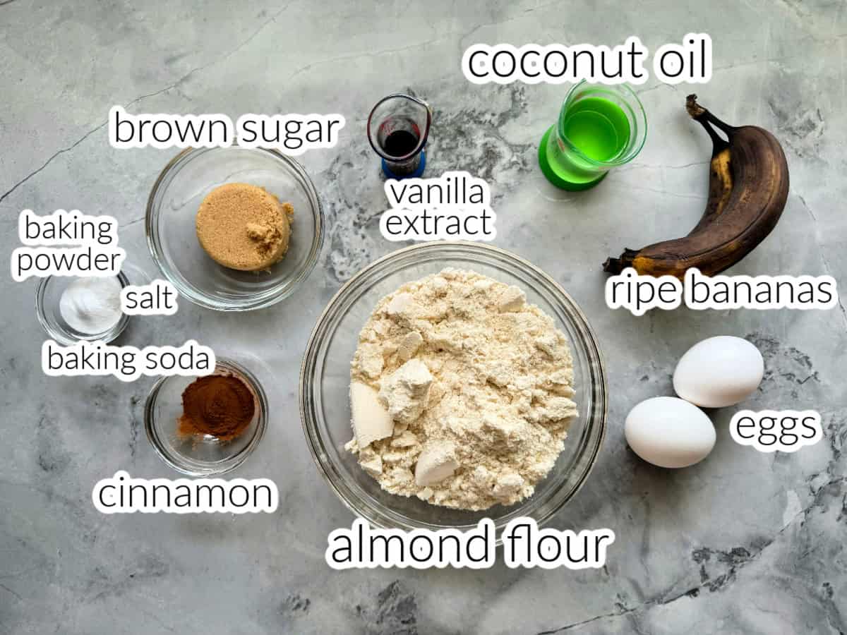Ingredients on counter: brown sugar, baking powder, salt, baking soda, cinnamon, almond flour, eggs, ripe bananas, coconut oil, and vanilla extract.