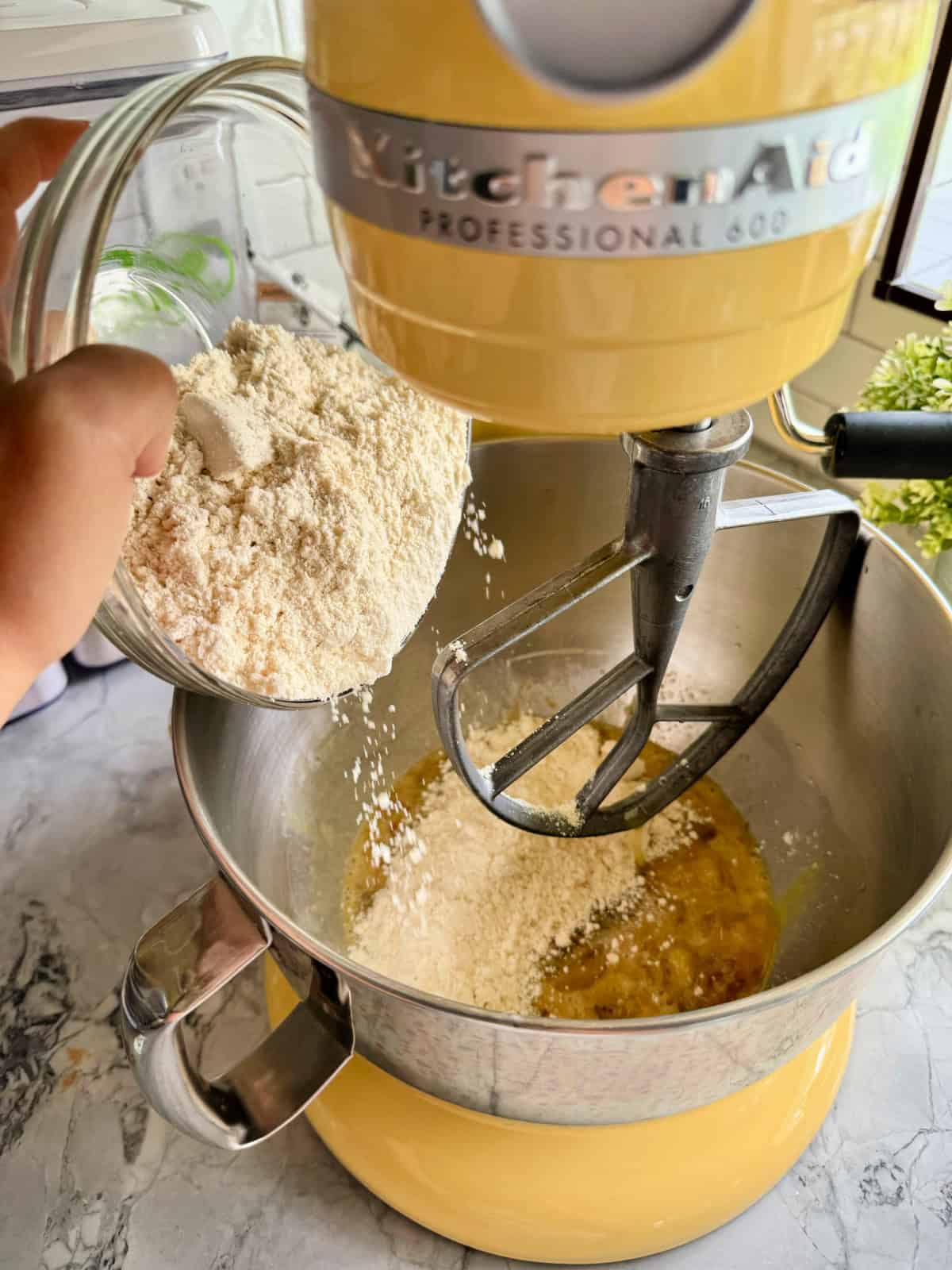 Hand pouring almond flour into a yellow KitchenAid stand mixer.