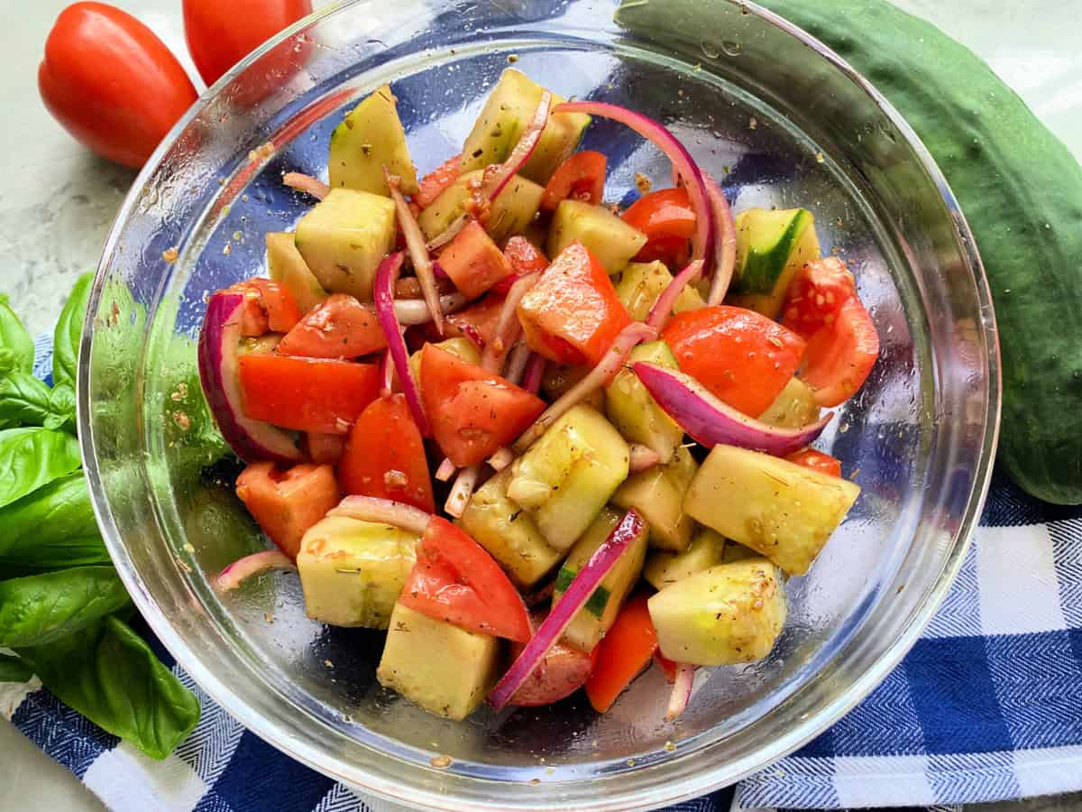 Glass bowl with cucumbers, tomatoes, onions mixed together with fresh produce next to it.