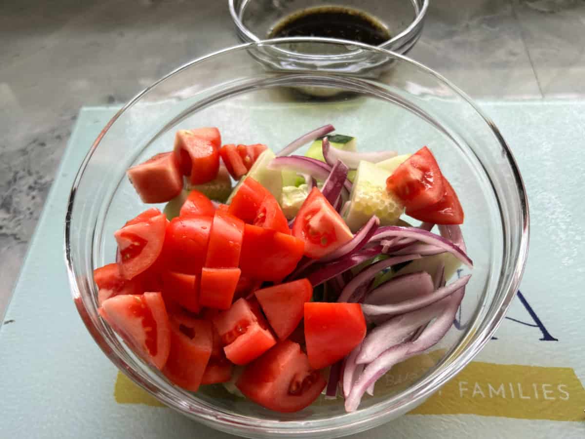 Glass bowl with cucumbers, tomatoes, onions, and a bowl of brown dressing in the background.