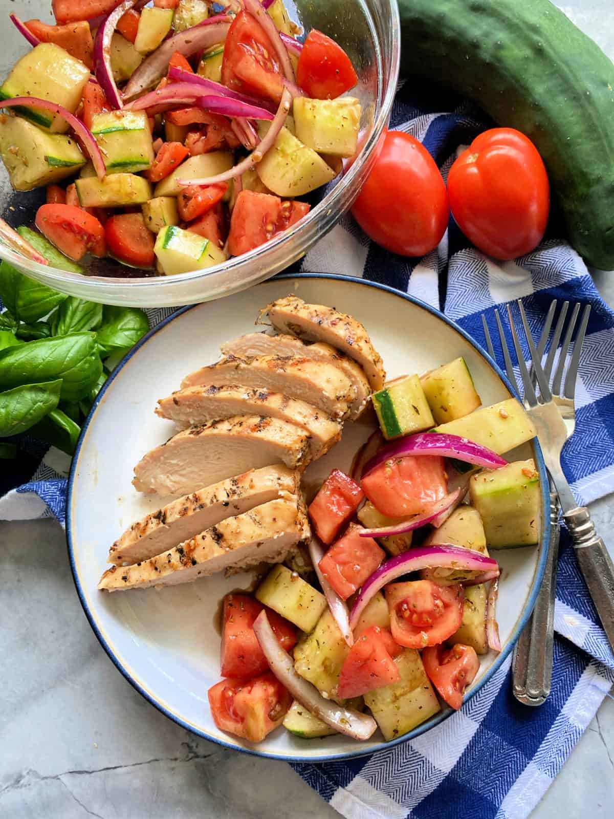 Plate filled with sliced chicken, veggie salad on a blue and white checkered napkin with a bowl of veggie salad in the background.