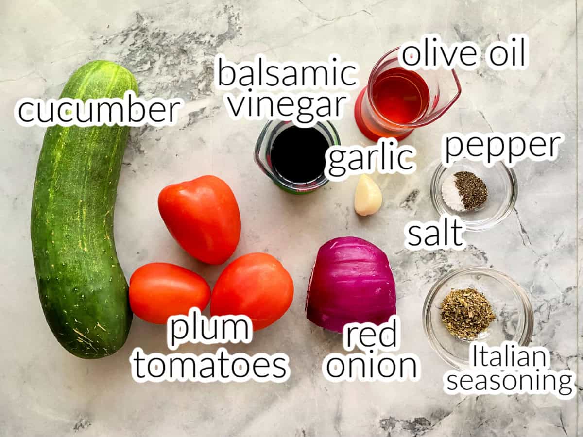 Ingredients on marble countertop: cucumber, tomatoes, red onion, salt, pepper, Italian seasoning, garlic, and balsamic vinegar.