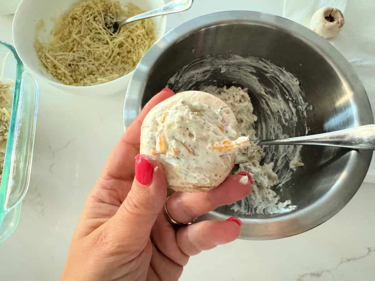 Female hand with pink nails holding a raw stuffed mushrooms filled with a cream cheese and cheese mixture.