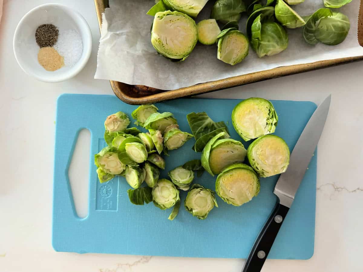 White countertop, blue cutting board with sliced brussels srpouts and a knife, with seasoning in a dish.