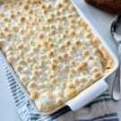 White baking dish filled with toasted marshmallow casserole on a white and blue striped cloth with a silver spoon next to it.