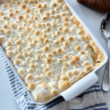 White baking dish filled with toasted marshmallow casserole on a white and blue striped cloth with a silver spoon next to it.