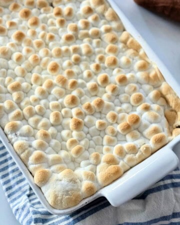 White baking dish filled with toasted marshmallow casserole on a white and blue striped cloth with a silver spoon next to it.