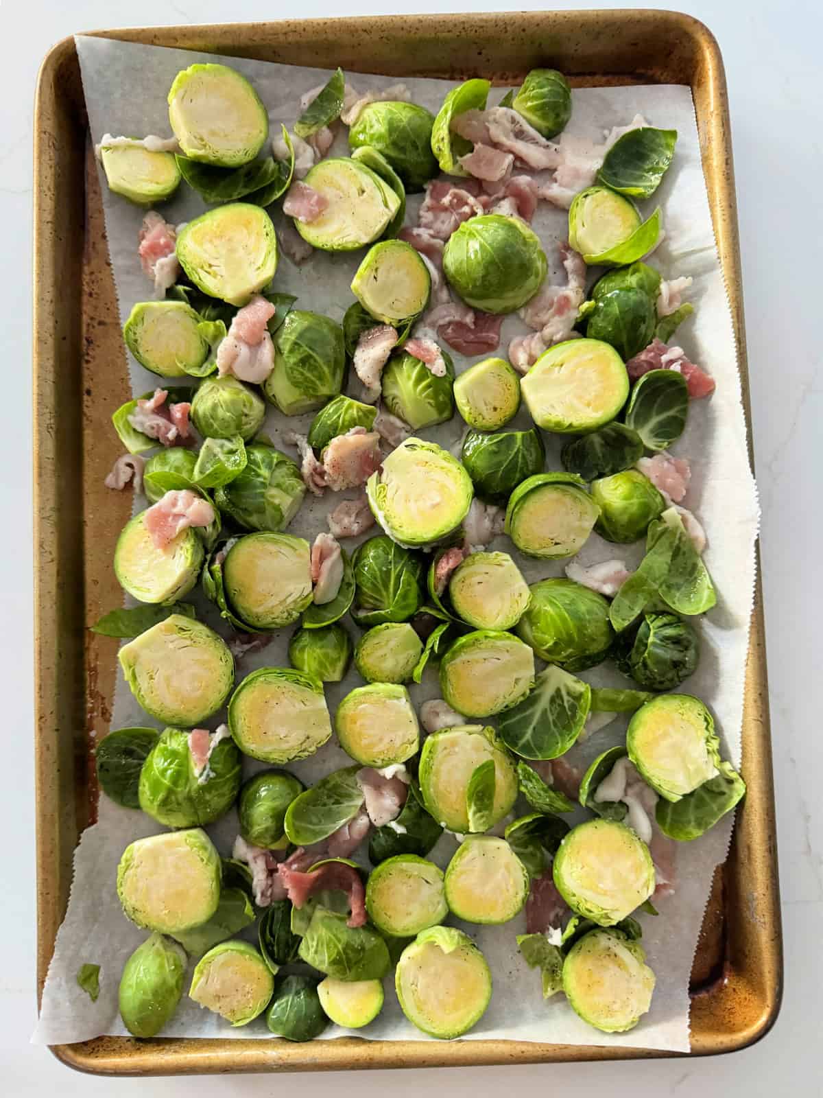 White counter with a parchment liner on a gold baking sheet with sliced brussels sprouts and bacon.