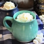 Two mugs with whipped cream with mini marshmallows on blue and white checkered cloth.