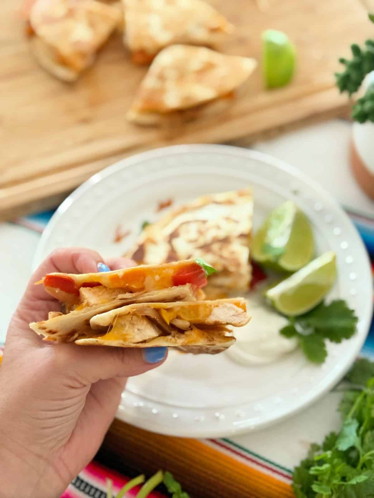 Female hand with blue nails holding 2 slices of quesadilla together over a white plate.