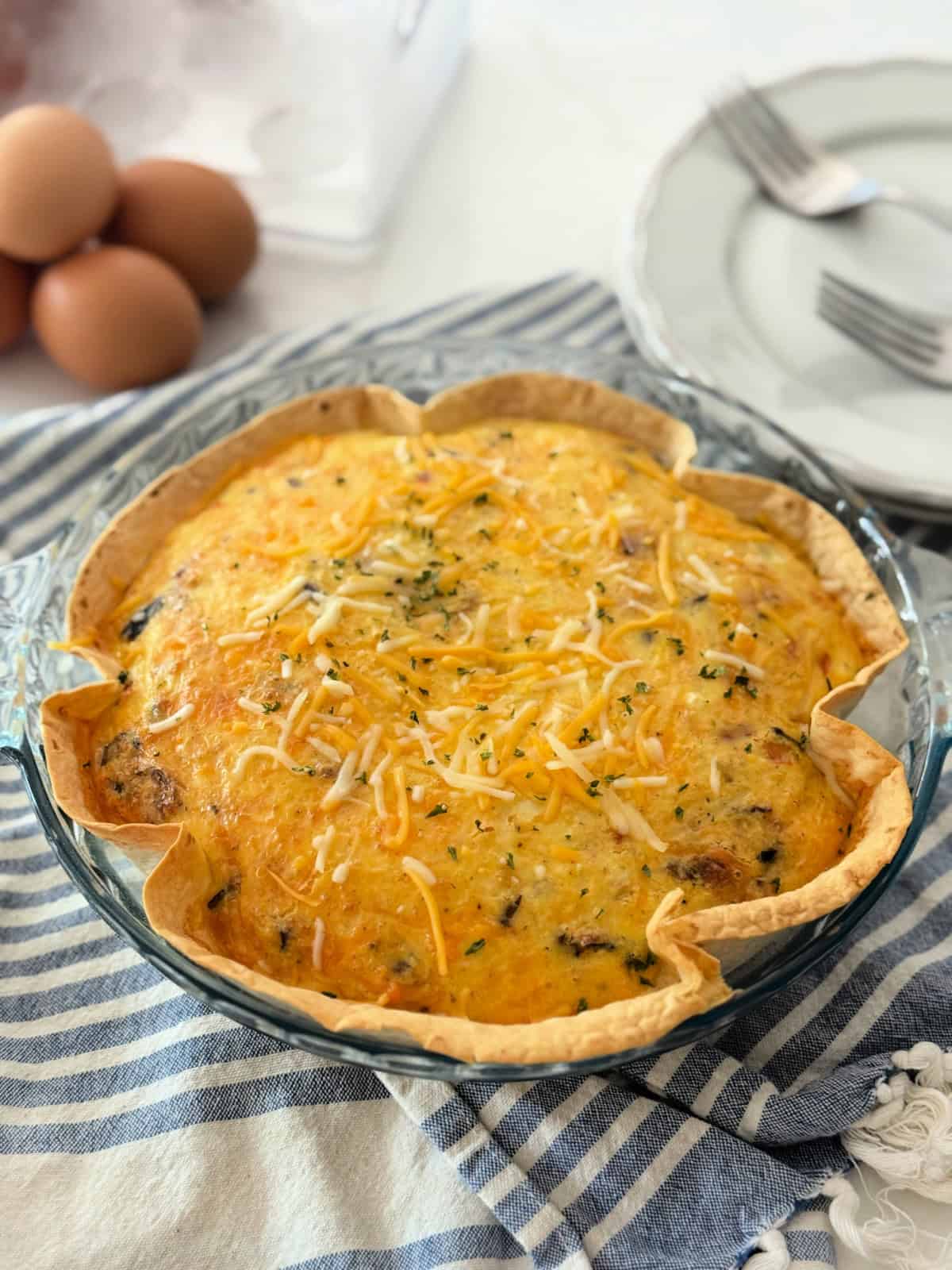 Glass baking dish filled with tortilla and egg topped with cheese resting on a blue and white cloth with brown eggs and plates in the background.