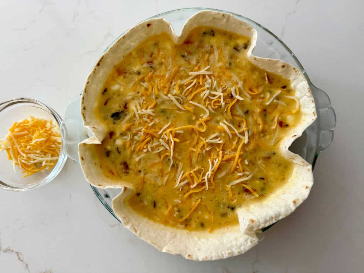 Tortilla in glass pie plate filled with egg mixture and shredded cheese on a white countertop.