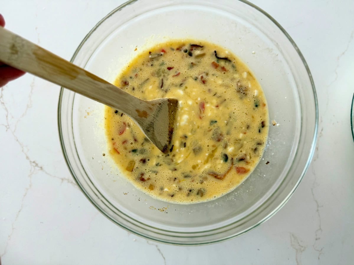 Clear glass bowl filled with scrambled eggs, vegetables, and cottage cheese with a wooden spoon in the mix.