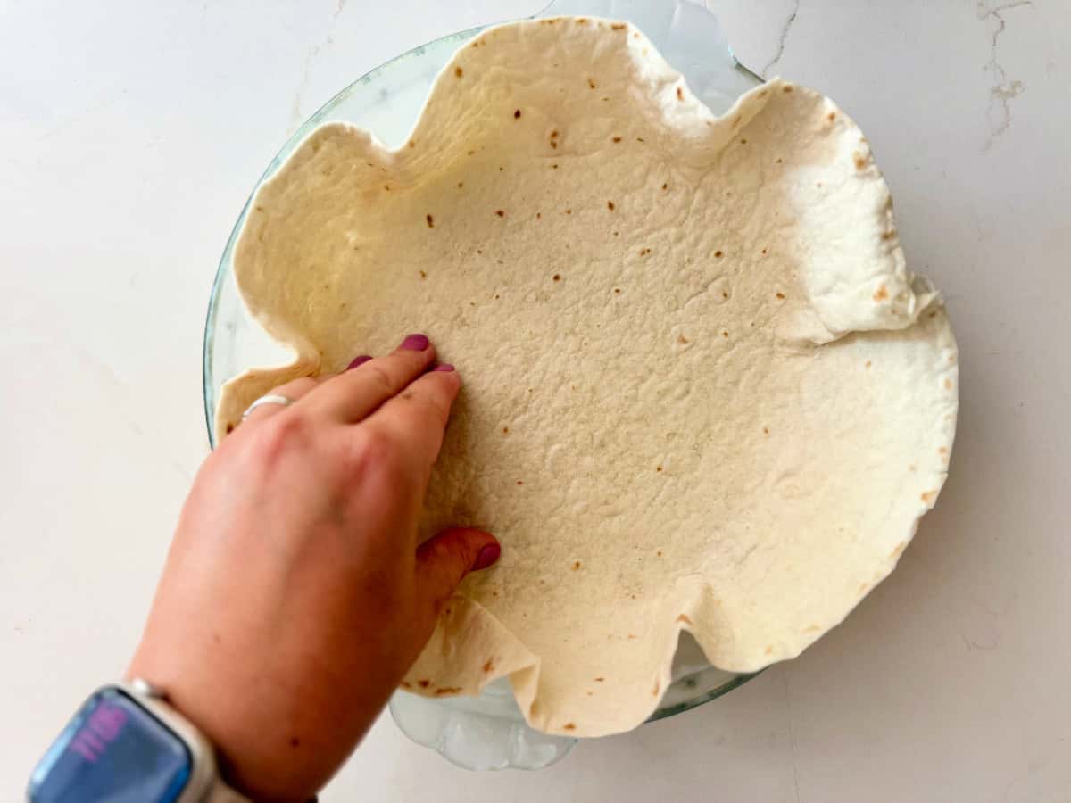Female hand with pink nails pushing down a tortilla in a pie plate on a white countertop.