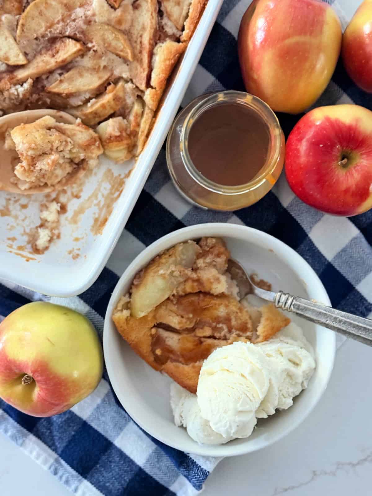 White bowl filled with apple cobbler and ice cream with caramel sauce, apples, and cobbler dish around it.