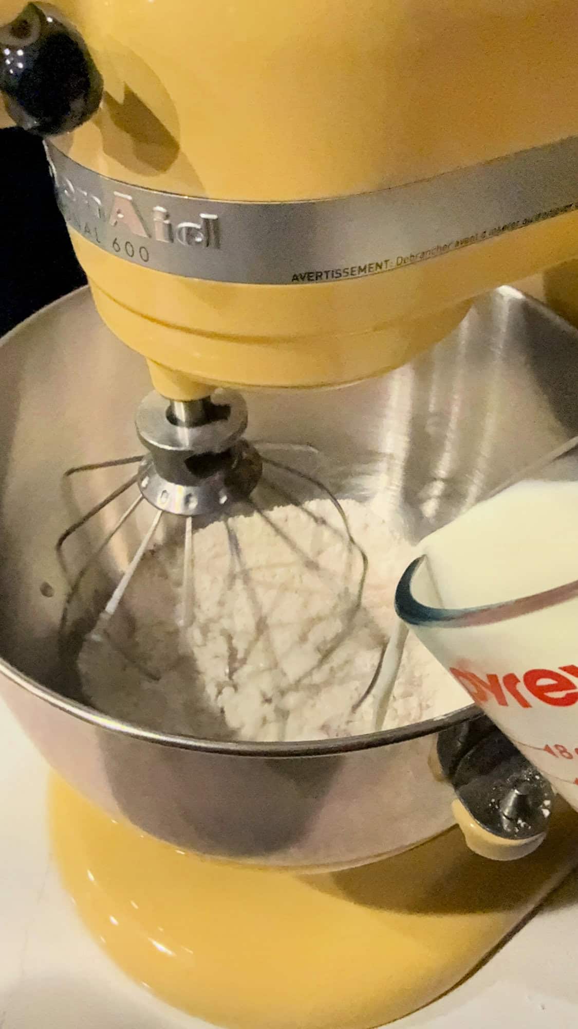 Yellow KitchenAid mixer fitted with whisk attachment mixing dry ingredients as milk is being poured into the bowl.