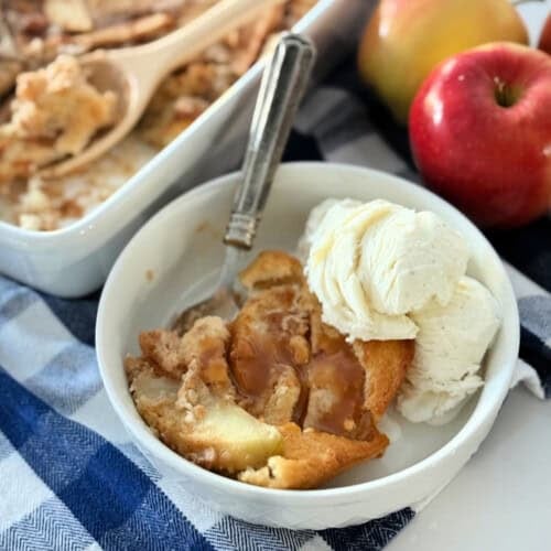 White bowl filled with apple cobbler, ice cream and a metal spoon.