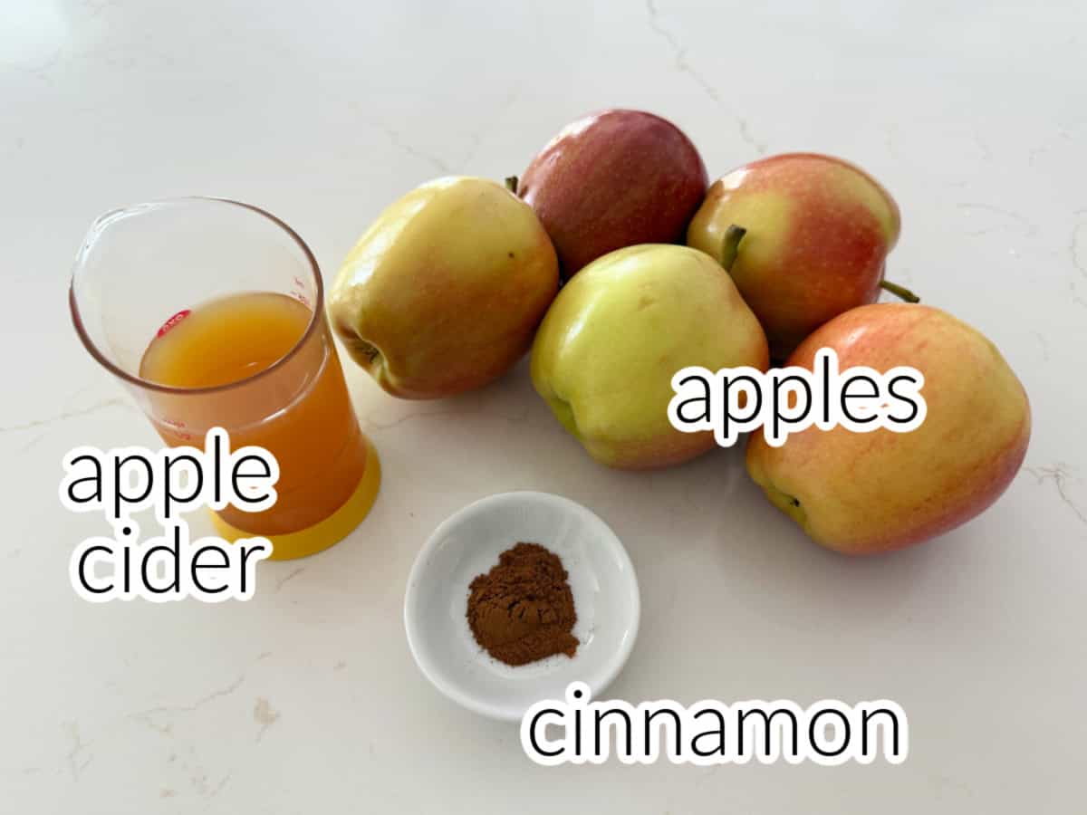 Ingredients on a white marble counter: apples, apple cider, and cinnamon.