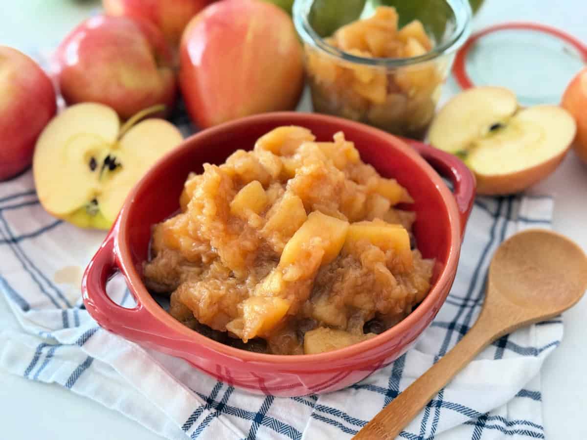 Red bowl with handles filled with chunky apple sauce on top of a white and blue striped cloth with a wooden spoon in front.