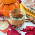 Glass jar with an orange spoon scooping out the brown spice with pumpkins and pie in background.