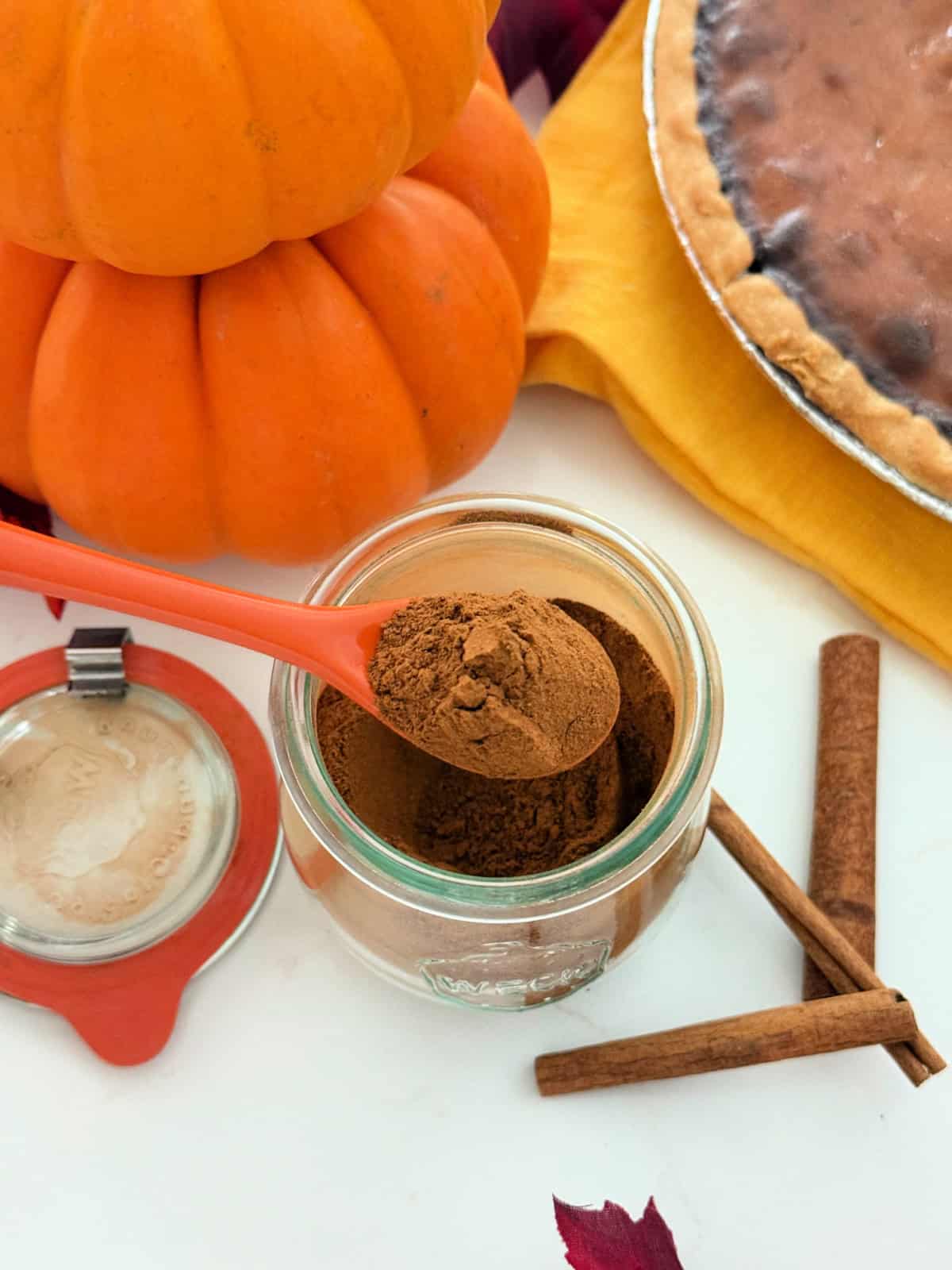 Glass jar filled with brown spices on an orange spoon with pie and pumpkins around it.