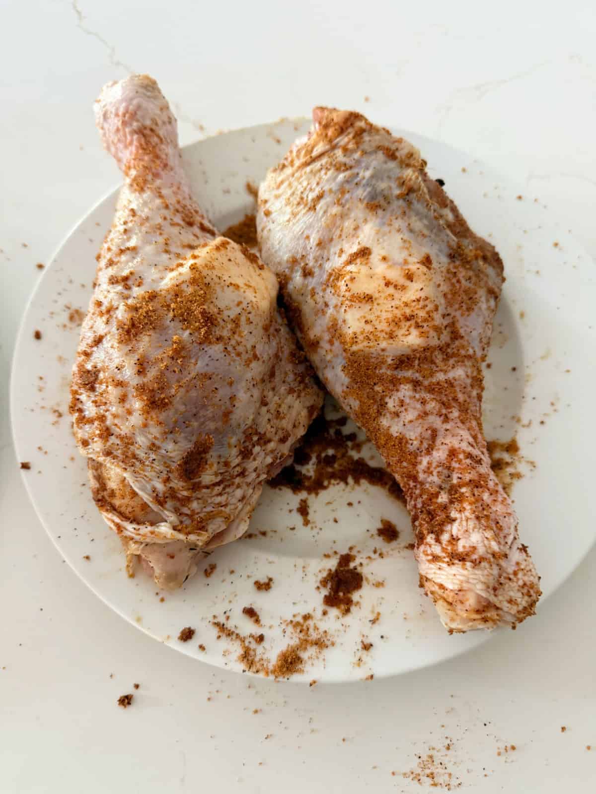 Two raw turkey legs with rub and butter under the skin on a white plate.