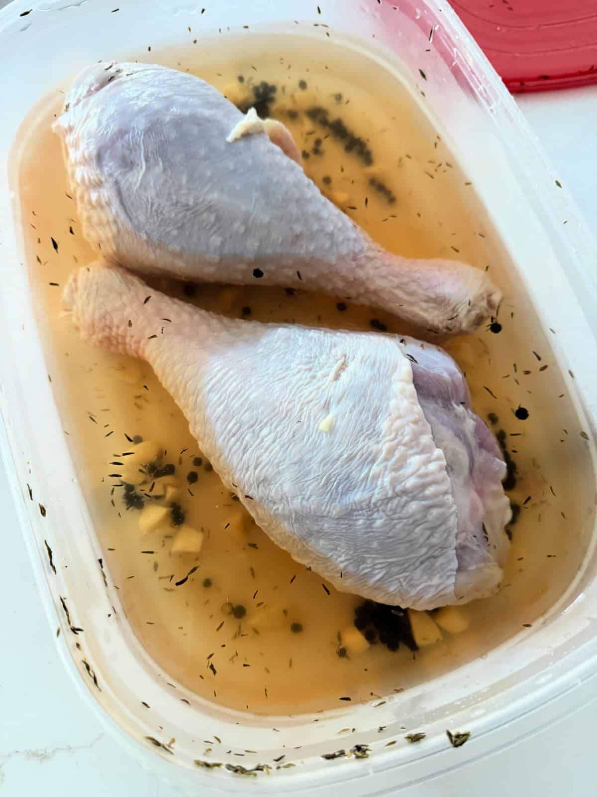 Two turkey legs in a plastic container sitting in a brine with peppercorns.