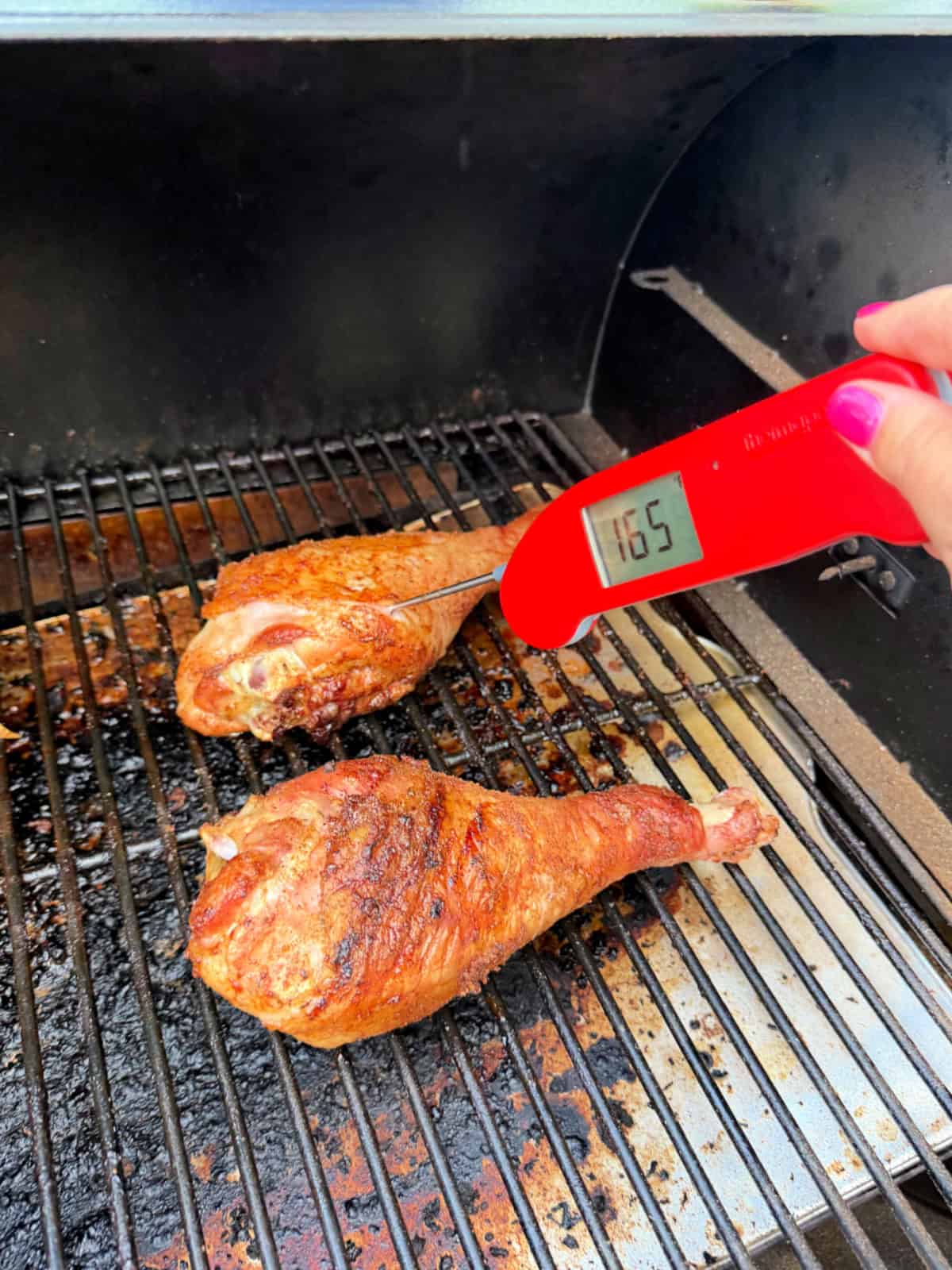 Female hand with pink nails using a red thermometer in a turkey leg on the grill.