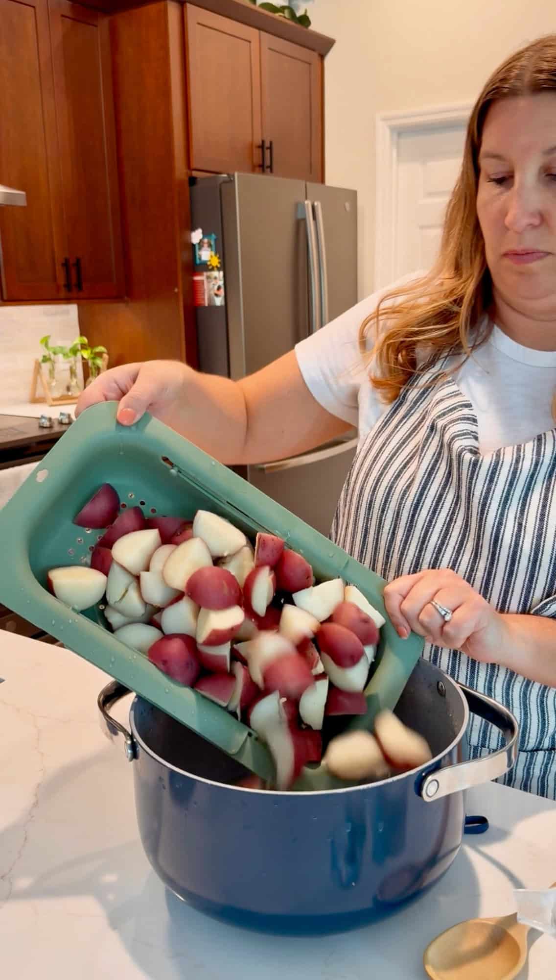 Female pouring quartered red potatoes from a green collander into a blue pot.