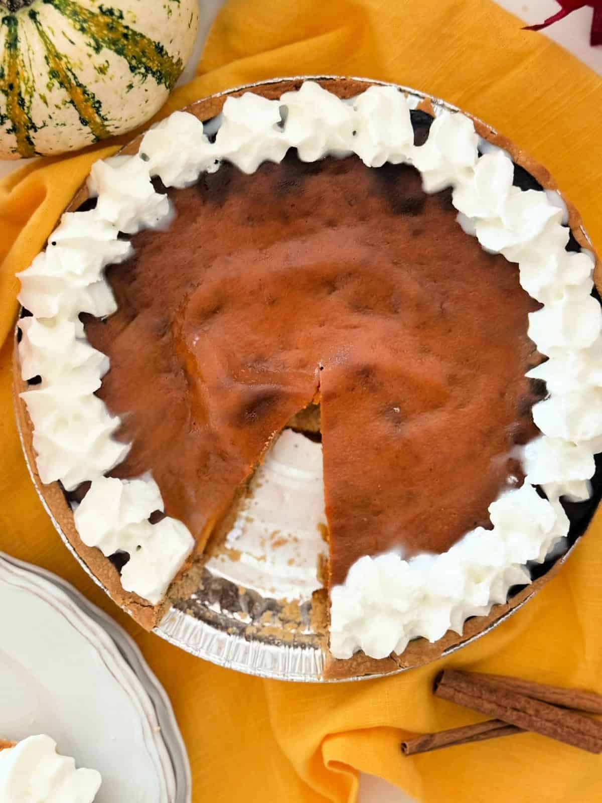 Pumpkin pie with whipped cream boarder with a slice taken out of it and cinnamon sticks next to it.