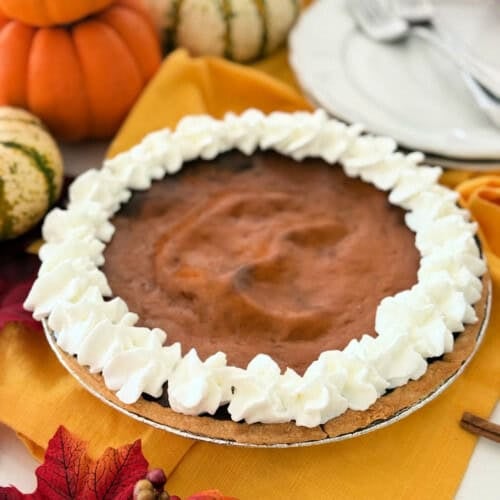 Pumpkin pie with piped whipped cream boarder resting on a yellow cloth with pumpkins and leaves around it.